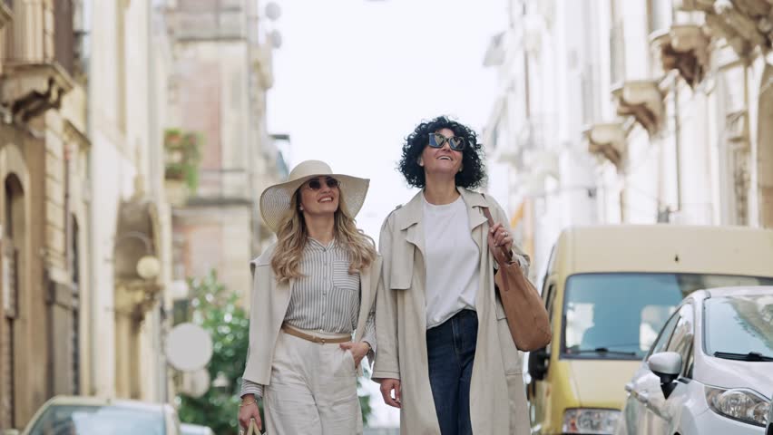 Two fashionable friends confidently stroll through a picturesque street in Catania, Sicily. They admire the historic architecture and vibrant city life, sharing joyful moments while exploring the charming Italian town.-