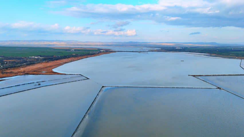 Aerial view of salt evaporation ponds under blue sky. Drone shot of geometric salt pans and coastal lagoons near green fields.