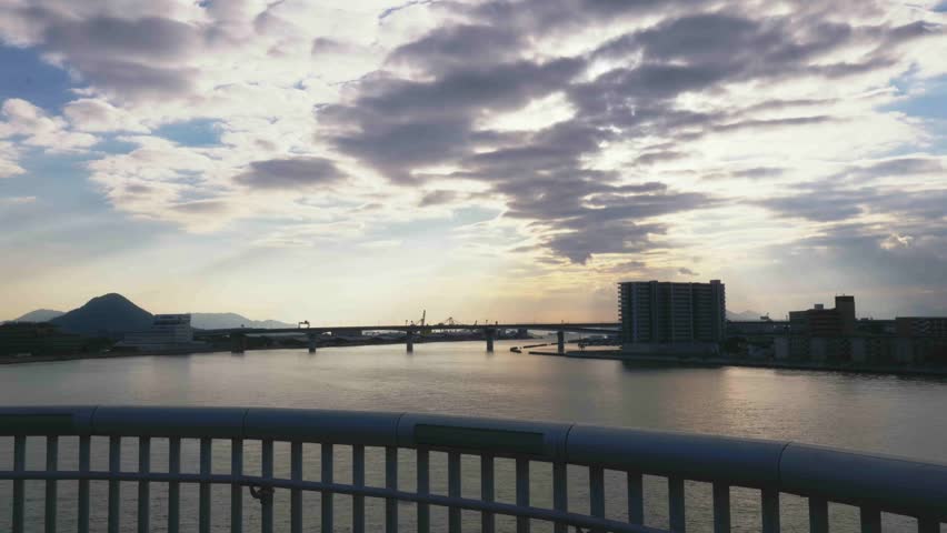 View of a wide urban river and coastal city at sunset from a bridge with a modern railing in the foreground. Dramatic cloudy sky with soft sunlight over the water and distant mountains.