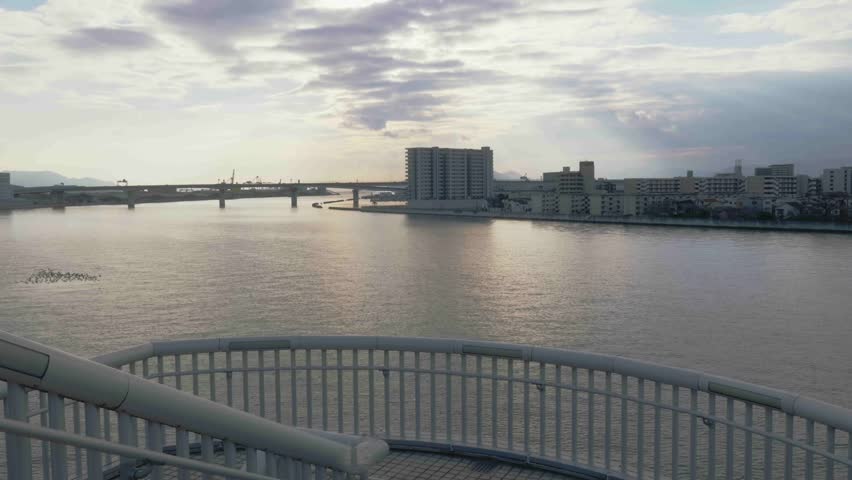 View of a wide urban river and coastal city at sunset from a bridge with a modern railing in the foreground. Dramatic cloudy sky with soft sunlight over the water and distant mountains.