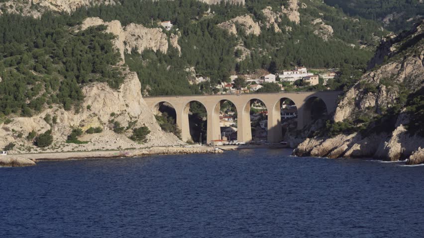 The iconic Marseille aqueduct bathed in the soft, golden light of sunrise. Majestic stone arches span the tranquil waters of the Calanques, surrounded by rugged cliffs and verdant hillsides, capturing the serene beauty of the French coast.