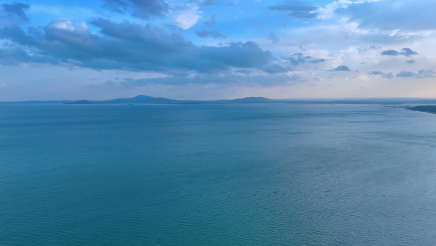 Wide seascape of calm blue ocean under cloudy sky at dusk. Panoramic view of a vast blue sea with distant hills and a soft cloudy sky during evening light.