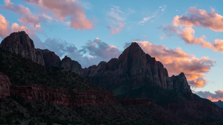 footage of the iconic The Watchman mountain peak and the surrounding Zion Canyon landscape at sunset. The sky is filled with dramatic clouds illuminated by the setting sun, creating warm pink and oran