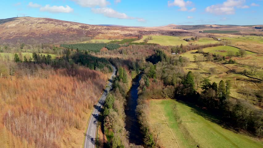 A road winds through green fields and dense trees in the Scottish Borders. This scene shows a clear view of the landscape near Langholm on the A7. Hills rise in the background under a blue sky