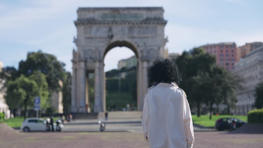 A woman carrying shopping bags leisurely walks towards the majestic Arco della Vittoria in Genoa, Italy. The historic piazza buzzes with daily life under a clear, bright sky.-