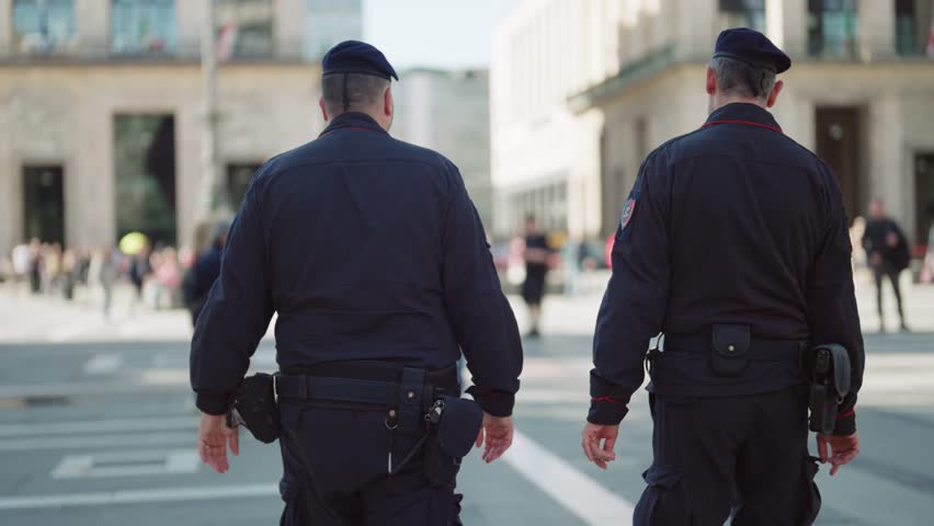 Two uniformed police officers maintain a visible presence, walking through a bustling public square in Milan. Their steady patrol ensures order and safety, reflecting urban security and community protection.