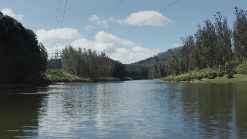 Drone pulls back over tranquil Sandynulla Lake near Sholur, Ooty, revealing forested hills and calm water surface.