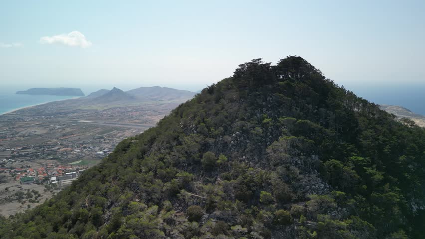 Aerial view of drone flying around Pico do Castelo mountain, with a shape of a vulcano. Porto Santo Island, Madeira, Portugal. Airport and long beach in the background