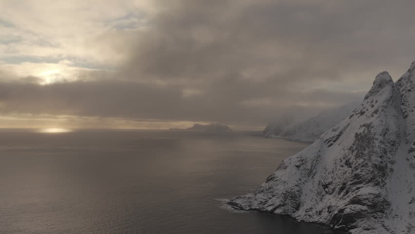 Cinematic aerial shot of snowcapped peaks meeting the dark arctic ocean under a moody winter sky in the Lofoten Islands, perfect for travel and nature documentaries.