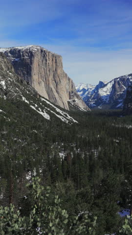 Vertical Screen: Yosemite National Park winter landscape featuring El Capitan granite cliff and Half Dome with snow-capped peaks, lush pine forest valley, wispy clouds and vibrant blue sky