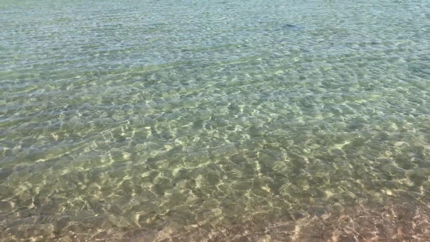 Clear shallow sea lapping rocky shore, transparent aquamarine water revealing pebbled seabed and granite boulders, sunlit ripples.