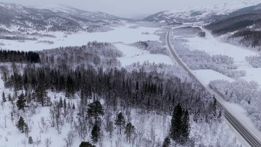 High angle aerial shot of a lone vehicle traveling along a cleared highway through a vast snowcovered forest and mountain landscape in the Lofoten archipelago.