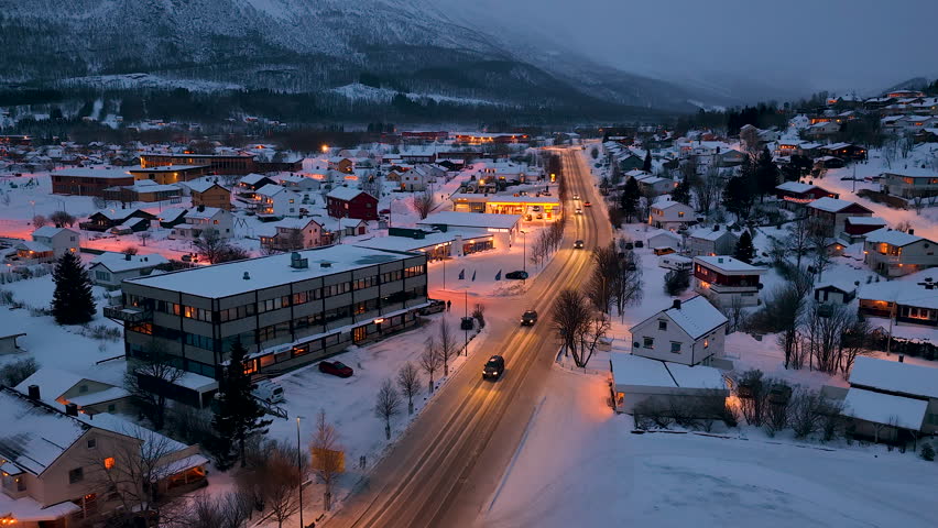 A cinematic aerial shot of a small Norwegian town in the Lofoten archipelago during a snowy winter twilight. Cars drive on illuminated roads past cozy houses and mountains.