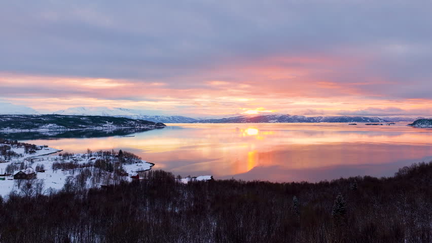 Breathtaking aerial view of a vibrant sunset reflecting on the calm waters of a Norwegian fjord during winter, featuring snowcovered mountains and coastal forests.
