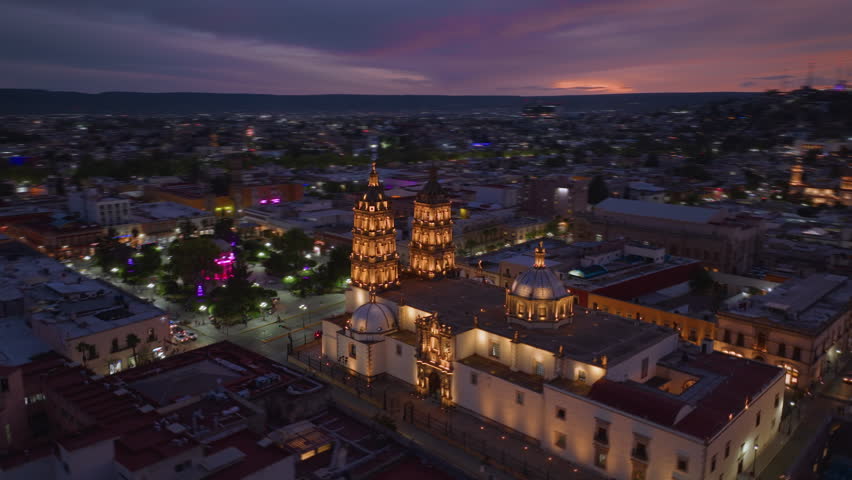 Aerial hyperlapse captures stunning Durango Cathedral bathed in golden lights at twilight in Durango, Mexico. Vibrant pink and purple sunset sky stretches across horizon as city lights twinkle below.