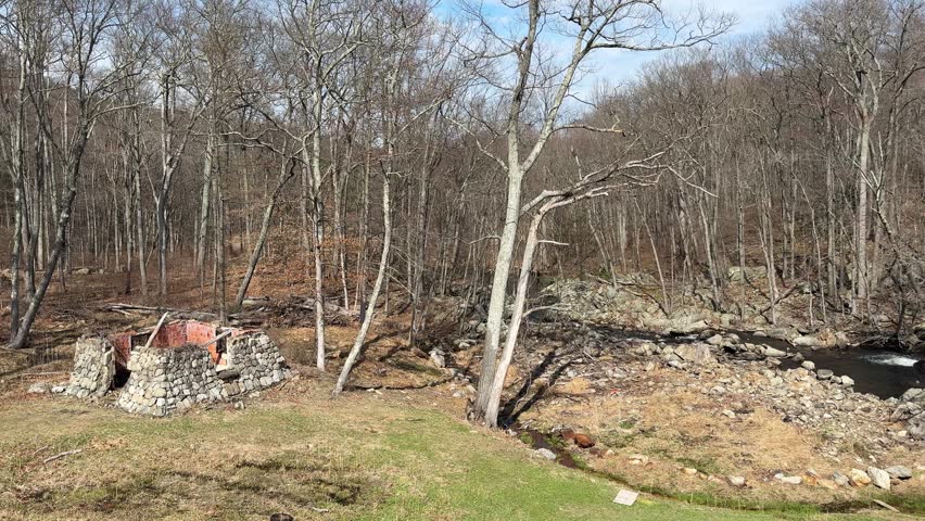 Old Stone house in Forest with Stream and Bare Trees. Rustic stone structure in a woodland forest surrounded by leafless trees and a small flowing stream. Natural outdoor landscape with early spring or late autumn atmosphere. Peaceful countryside scene with elements of history and nature. horizontal