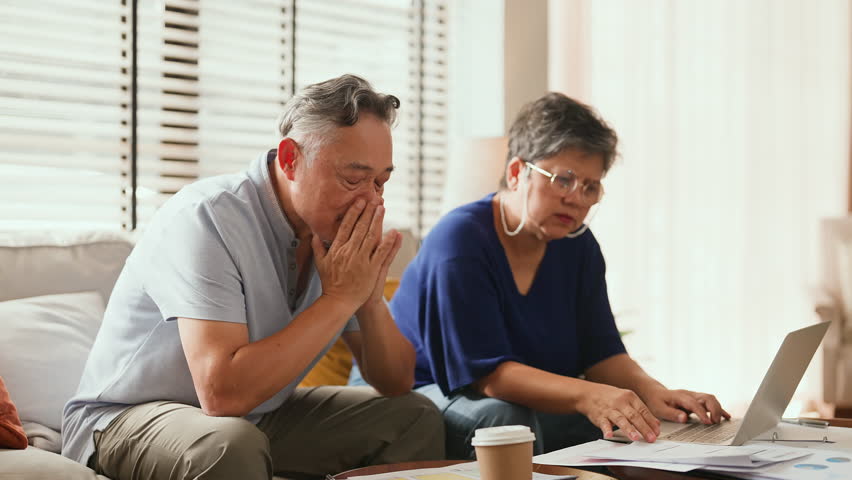 Unhappy Asian senior couple sitting on sofa at home facing financial problem, stress from debt and bills, worried about insurance and money, retirement struggle, emotional tension and family hardship.
