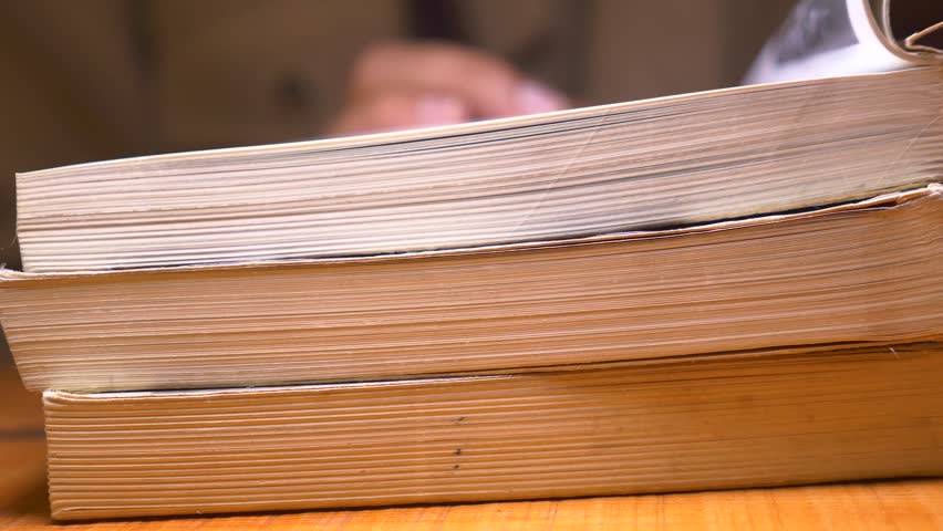 Footage shows stack of old books on wooden table, person in background flips through pages of another book or magazine.