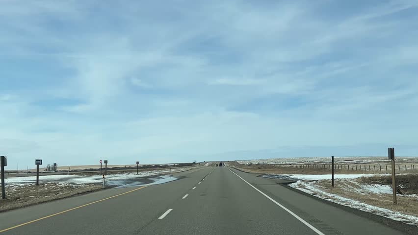Point of view driving footage along a rural highway through open prairie farmland with light snow coverage in Alberta, Canada. The scene features a straight road, wide landscape, and winter countryside environment under a blue sky.