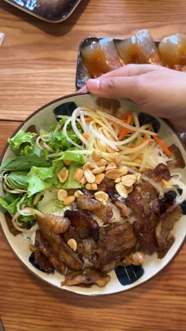 A person rotates a bowl of Vietnamese grilled pork rice noodles on a wooden table