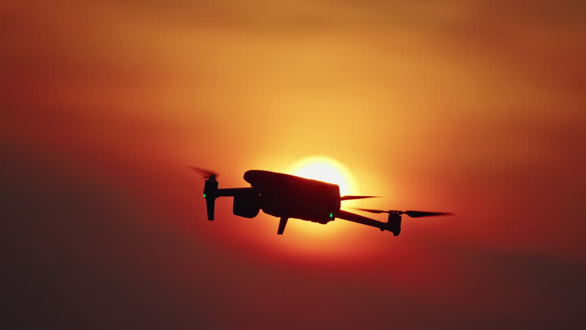Drone flying in front of glowing sun during sunset with dramatic orange sky.