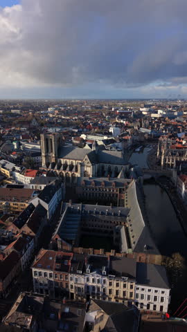 Vertical Screen: Aerial view of Ghent Belgium European city featuring Gothic cathedral, historic buildings with red rooftops, canal waterway, and dramatic cloudy sky over urban landscape
