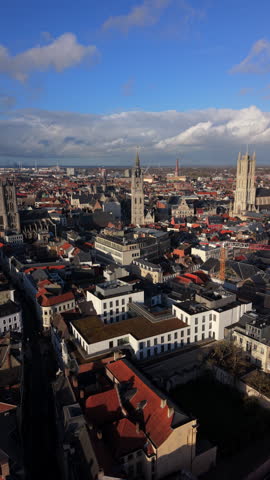 Vertical Screen: Aerial view of Ghent, Belgium featuring iconic medieval belfry and cathedral towers rising above traditional red-tiled rooftops under blue sky with clouds