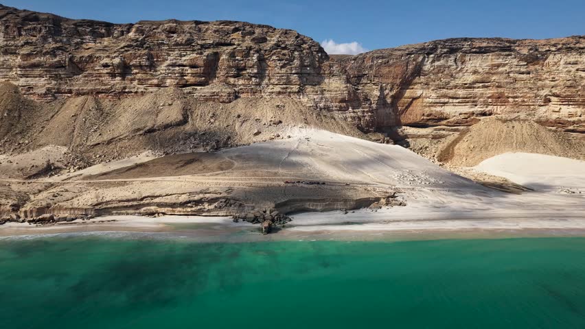 Aerial view of a rugged desert coastline with sandy beach, turquoise water, and layered rock cliffs; a dirt road with small vehicles runs along the shore under harsh sunlight.