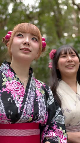Two Japanese women wearing floral kimonos smile and look upward in a lush green forest