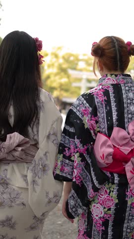 Back view of two women wearing floral kimonos walking through a sunny Japanese shrine garden
