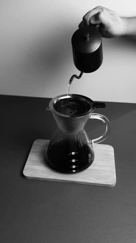 Black and White Pour Over Coffee Making, Artistic black and white close-up shot of a person pouring hot coffee into a cup in a minimalist setting.