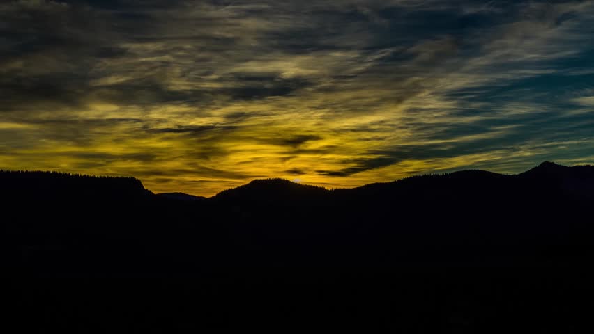 A stunning, vivid sunset illuminates the sky with yellow and orange clouds above a dark, silhouetted mountain range. This scenic landscape captures the dramatic beauty of nature at dusk or dawn.