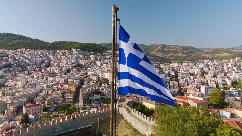 Aerial view of the Greek national flag waving proudly atop the central tower of the historic Kavala fortress. Stone walls, battlements, and the old town below, Kavala, Greece