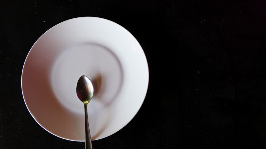 A single silver spoon rotating on a white plate over a dark background with empty space on the right, top down close-up shot