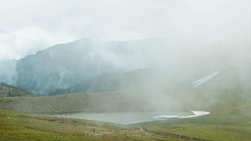 wide shot footage of a serene, fog-covered mountain landscape featuring a small, calm lake and lush green hillsides. The ethereal mist rolls over the rugged peaks, creating an atmospheric and tranquil