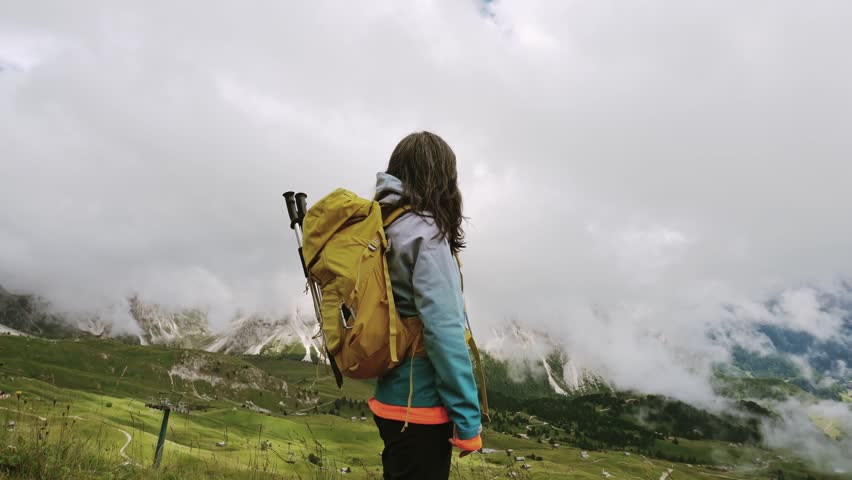 Female hiker with a yellow backpack overlooking the misty Dolomites, Italy. Dramatic mountain peaks, low clouds, and green alpine slopes. Cinematic travel B-roll. Trail trek Dolomiti Italy