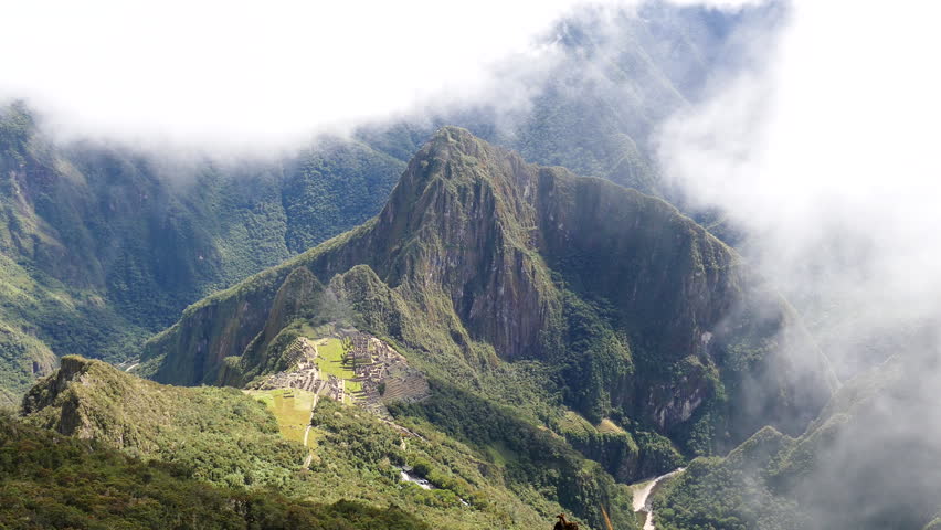 Machu Picchu Ruins with Huayna Picchu and the Urubamba River in Rising Mist, Peru. A static aerial view from Machu Picchu Mountain, showing the ancient Inca citadel, Huayna Picchu, and the Urubamba River Valley in Peru. Low clouds drifting through green mountain landscape create dramatic atmospheric mood. This iconic UNESCO World Heritage site in the Andes Mountains is a popular tourist destination.