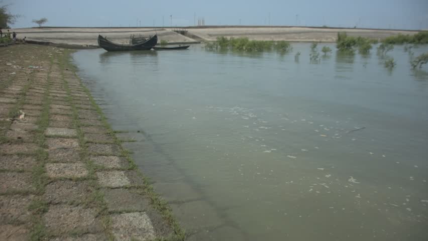 Tidal surge from the Bakkhali River pushes water up to the town’s flood-protection embankment, showing wave impact on the structure and nearby shoreline under natural light.