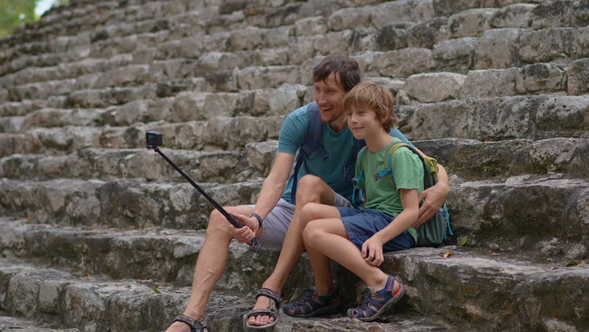 A young man and his son, tourists, are exploring ancient Maya ruins in Latin America. They are taking a selfie while sitting on the stone steps of a temple.