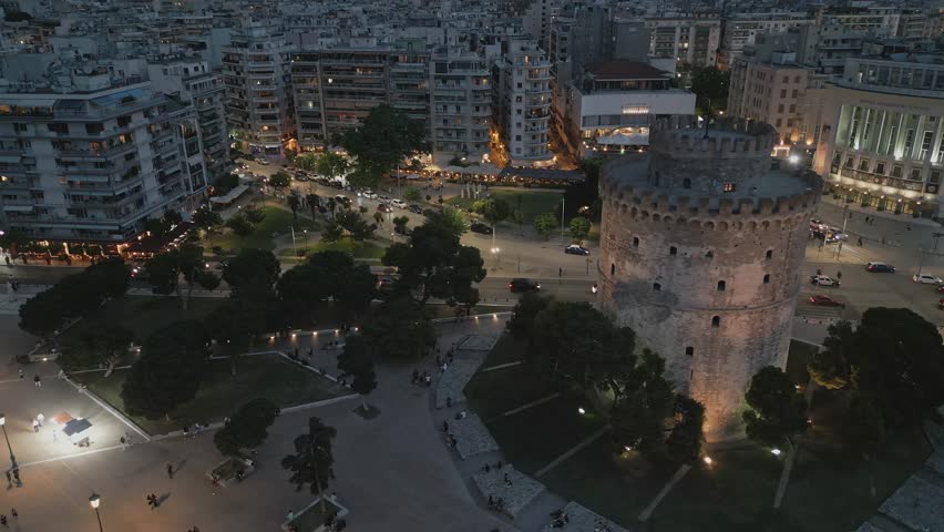 Cinematic aerial orbit shot around the illuminated White Tower of Thessaloniki at night. City lights, traffic, and park view, Greece.