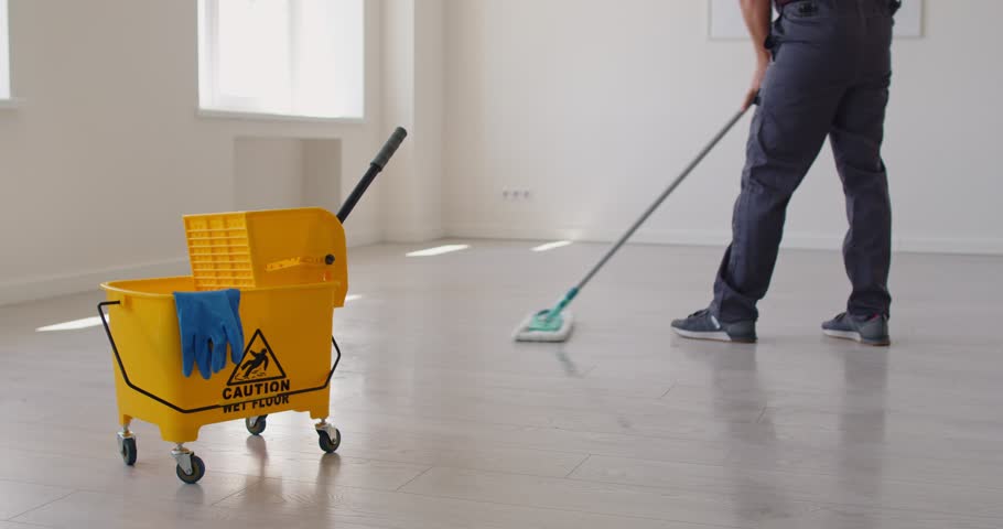 Close-up shot of an male cleaner mopping floor near yellow caution sign, providing housekeeping service, handling mop for cleanup, ensuring thorough cleaning with attention to safety.