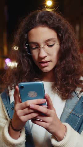Portrait of happy smiling teenager girl in eyeglasses using smartphone typing text messages browsing internet social media web app chatting online. Young tourist in city street at night. Lifestyles