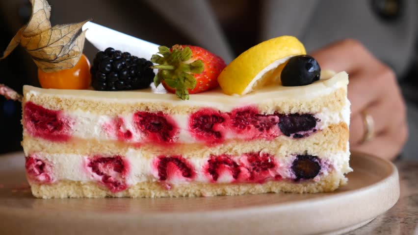 Close up view of hand cutting into sweet dessert with fork, revealing layers of cake, cream, and vibrant berries.