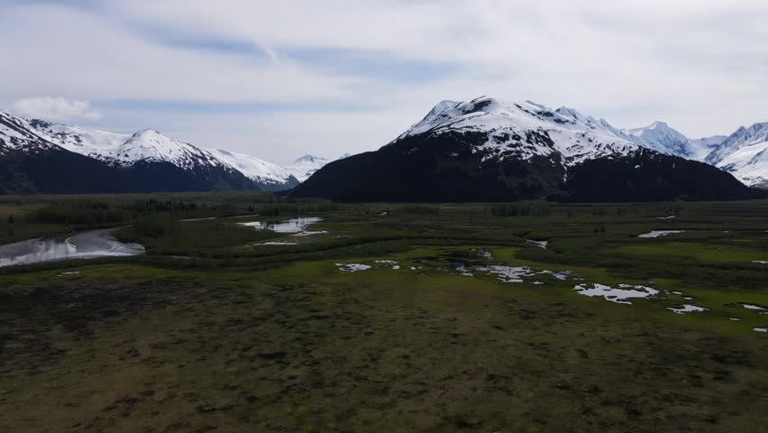 Aerial drone footage of Alaska’s forested mountains with snow-capped peaks and a winding river below, cinematic scenic wilderness landscape.