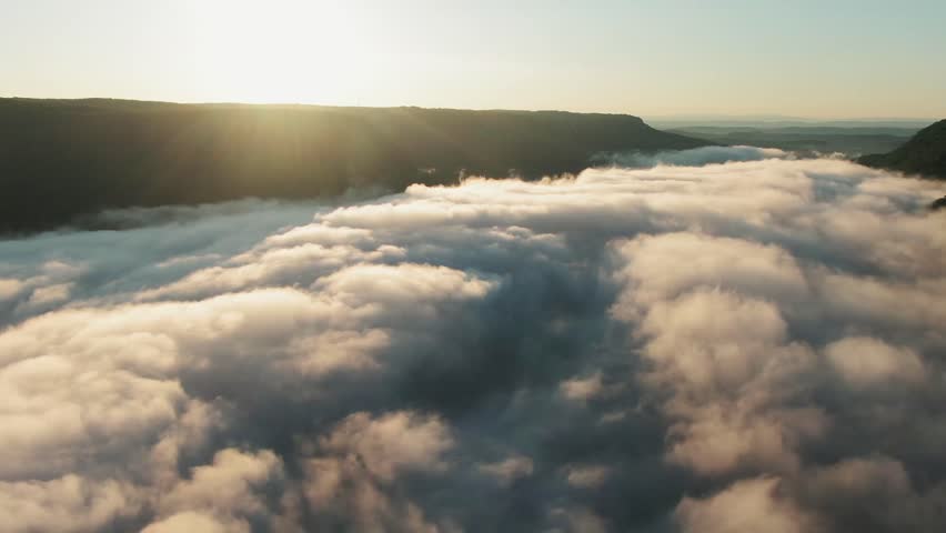 Aerial landscape of clouds drifting over hills during sunrise.
