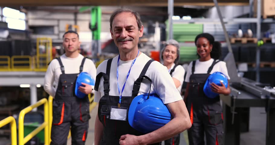 Senior male leader smiling at the camera holding a blue hard hat with his multi ethnic team of factory workers standing confidently in the background inside an industrial warehouse facility