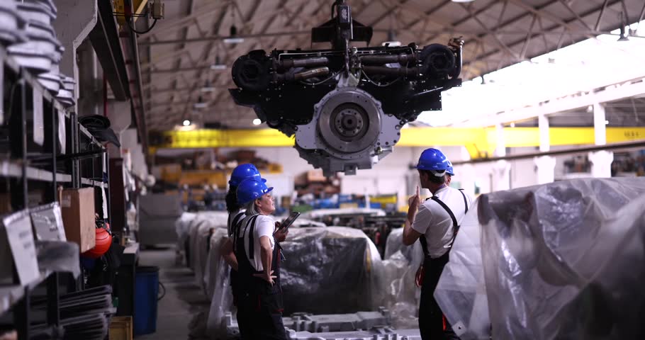 Industrial engineers in hard hats supervising the assembly of a large diesel engine hanging from a crane in a heavy industry manufacturing plant, discussing production and logistics details
