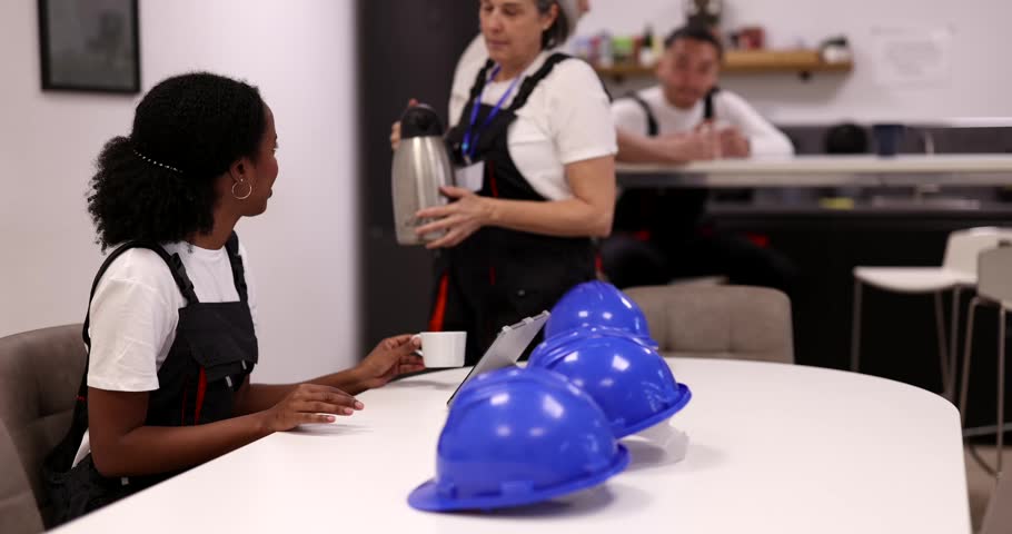 Two diverse female factory workers wearing overalls enjoy a coffee break together, with one woman pouring a hot beverage from a thermos for her colleague in the company canteen
