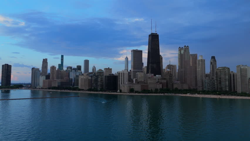 Aerial view of Chicago skyline along Lake Michigan waterfront during blue hour. Iconic skyscrapers including tall black tower rise against dramatic cloudy sky with boats on calm water below.