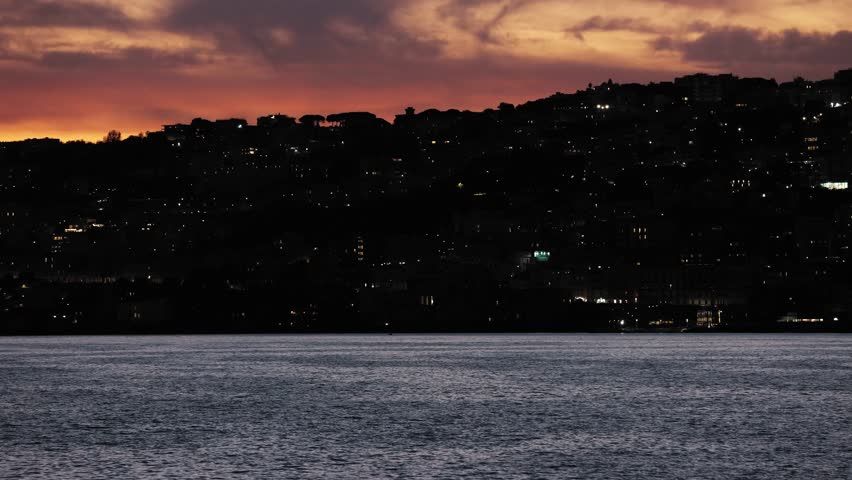 Naples city lights glow across the hillside during late twilight. The dark Mediterranean Sea reflects the fading sky as the urban coastline transitions into night. Useful for backgrounds.